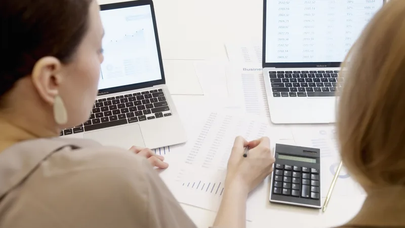 Two colleagues working through printed figures with a calculator beside two laptops