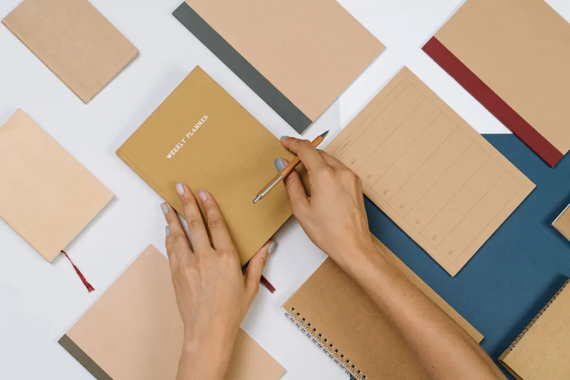 Hands holding a pencil over a weekly planner notebook on a desk