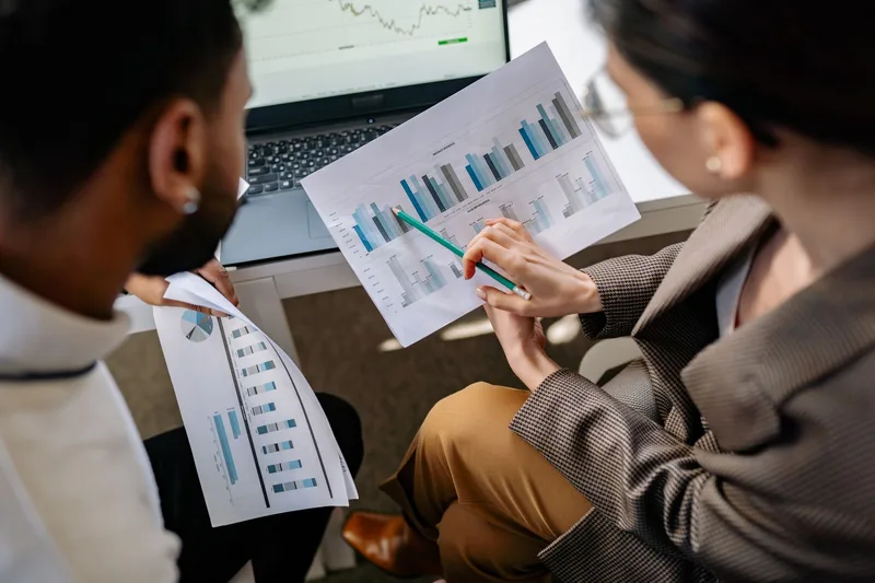 Two colleagues pointing at bar charts on printed reports beside a laptop